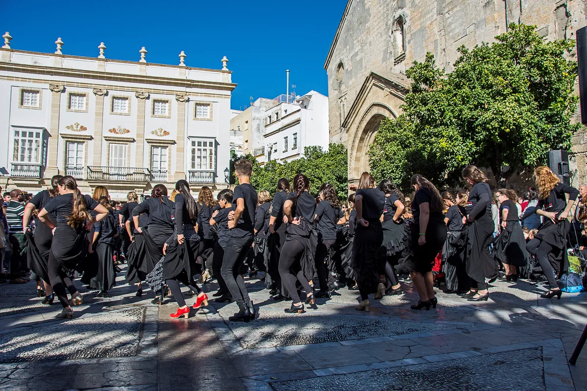 Eine große Gruppe von Tänzerinnen in schwarzen Röcken und Oberteilen, die gemeinsam auf einem sonnendurchfluteten Platz in Jerez auftreten, links ein weißes Stadthaus, rechts ein steinerner Kircheneingang