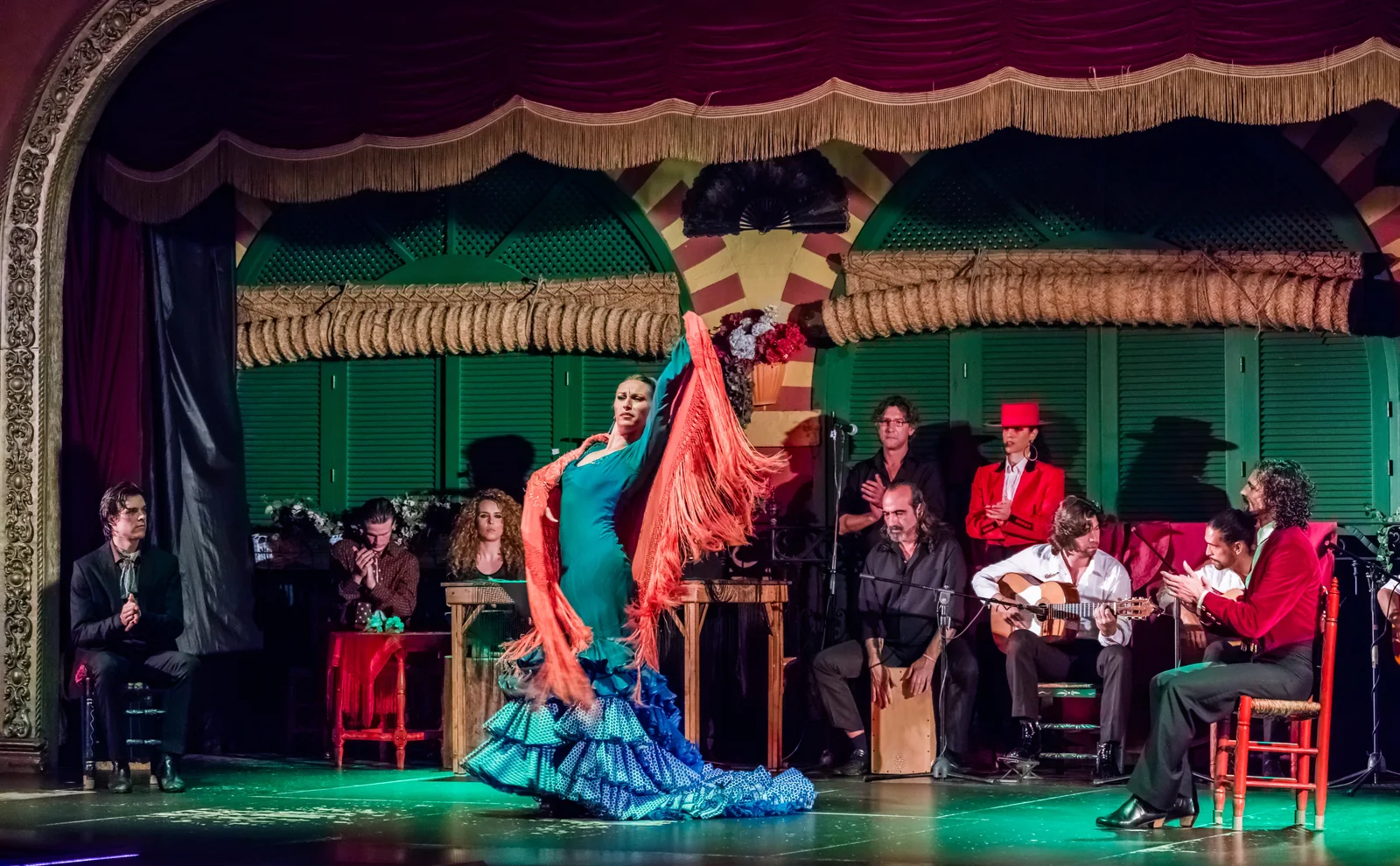 Tancerka flamenco w turkusowej sukni i pomarańczowej chustce z frędzlami wiruje na zielono oświetlonej scenie, a za nią, pod czerwoną welurową kurtyną i łukami podkowiastymi, siedzą gitarzyści, śpiewacy i palmeros